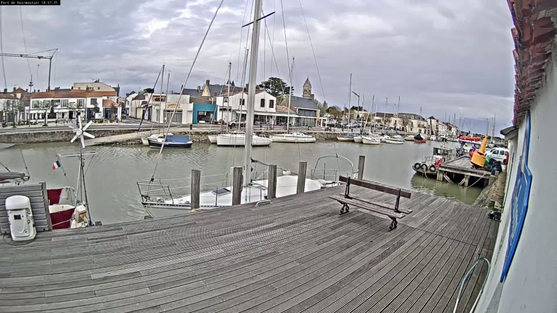 A harbor filled with sailboats and small motorboats under a cloudy sky, viewed from a wooden dock with a bench.