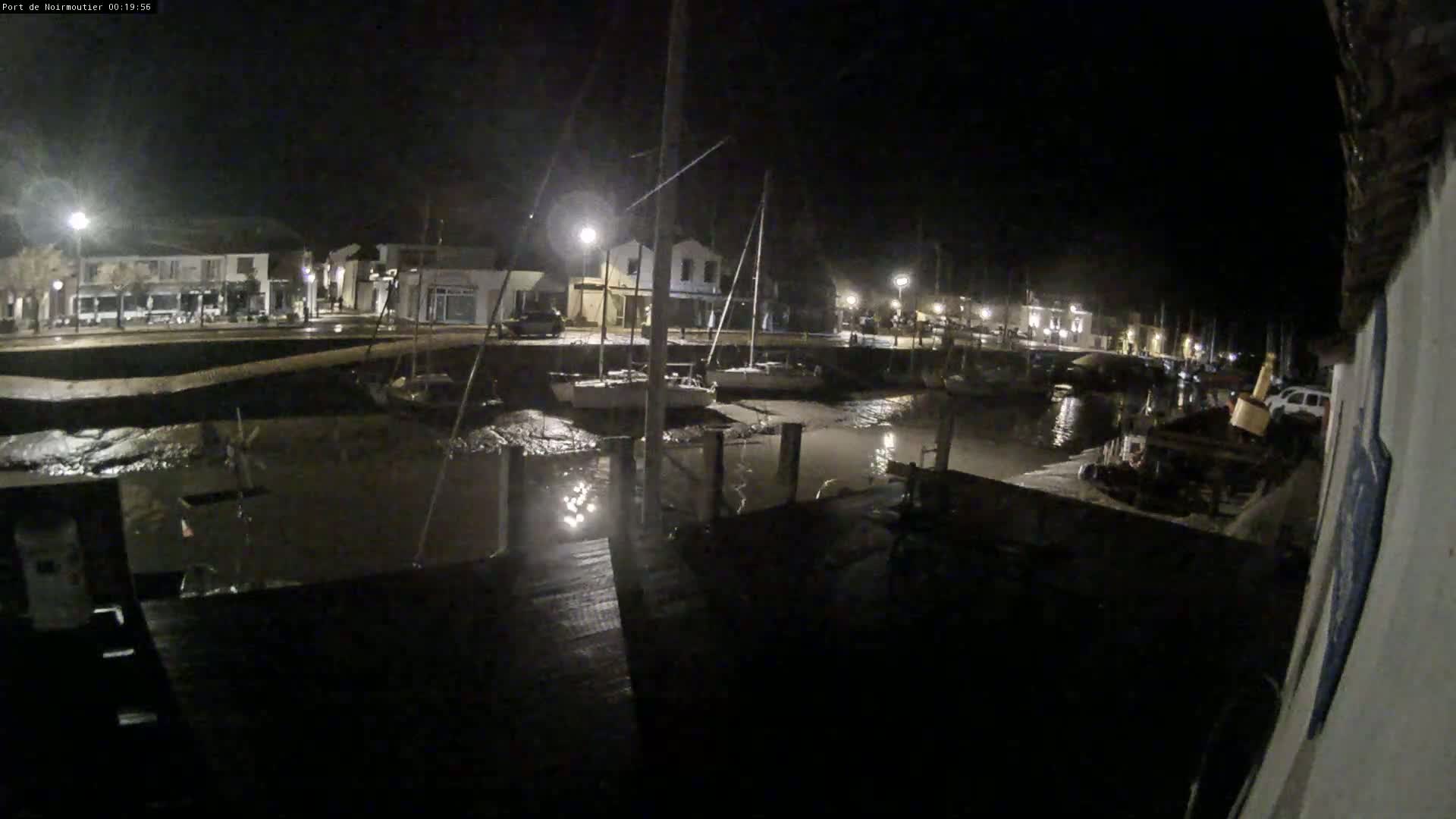 At night, a harbor is mostly empty except for several sailboats docked at low tide next to a waterfront town illuminated by streetlights.