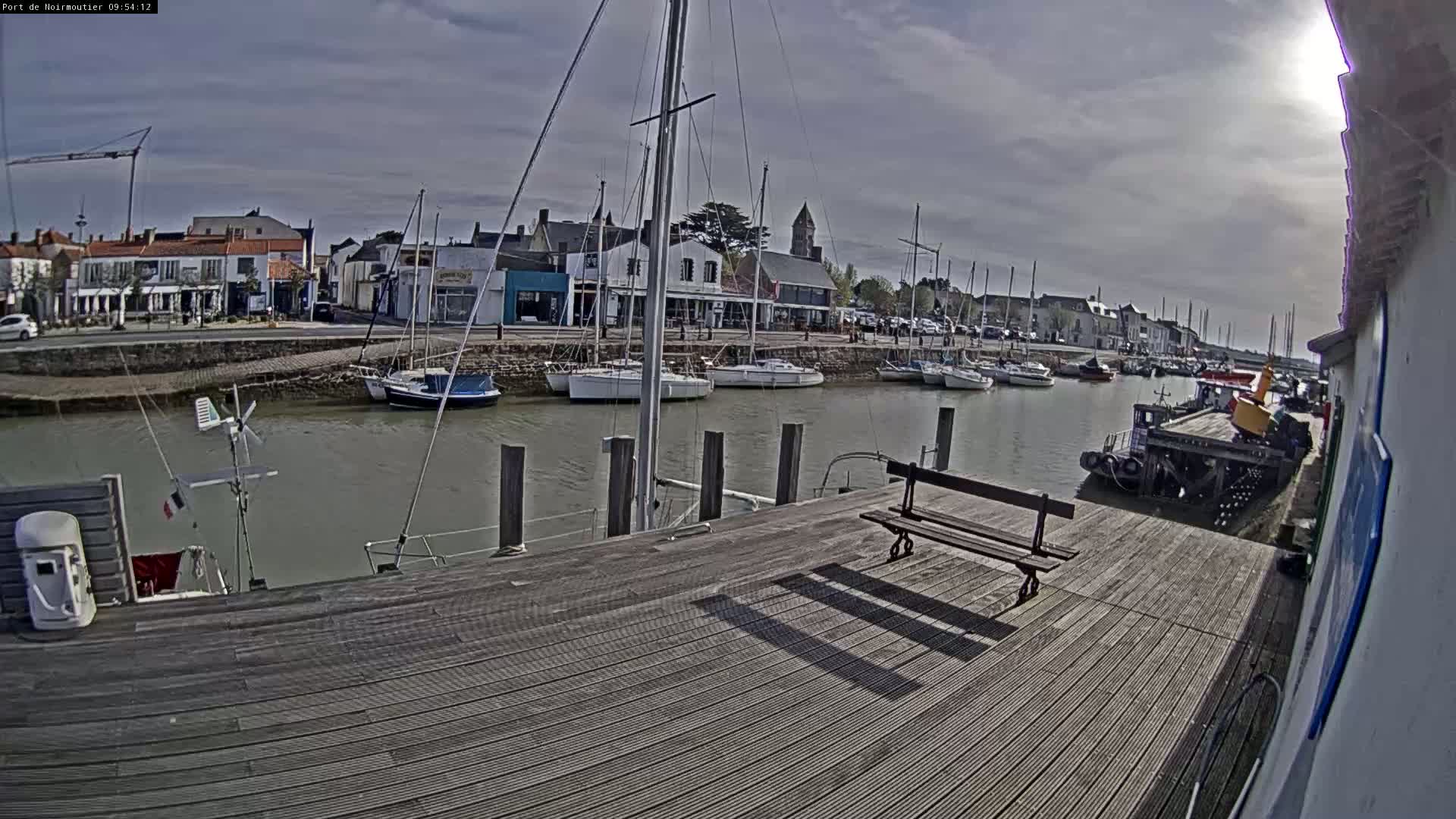 A wooden dock overlooks a calm harbor filled with sailboats and a tugboat, under a partly cloudy sky.