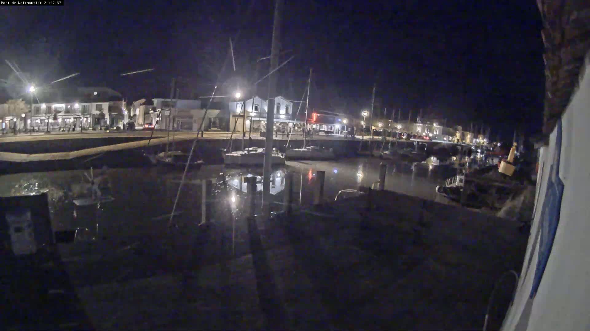 At night, a harbor filled with boats is visible, with calm water reflecting the lights from buildings along the waterfront.