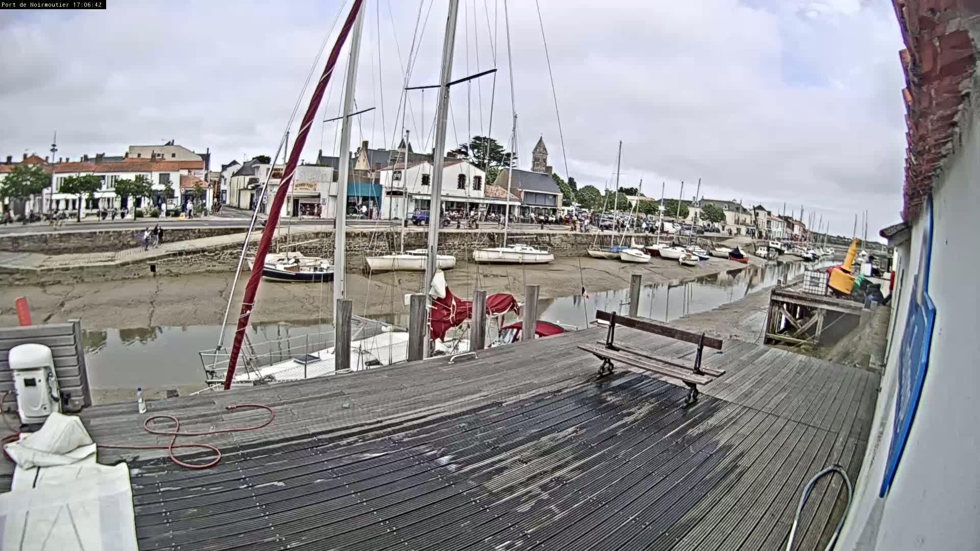 A wooden dock overlooks a harbor at low tide, with numerous sailboats moored and a town visible in the background under a cloudy sky.