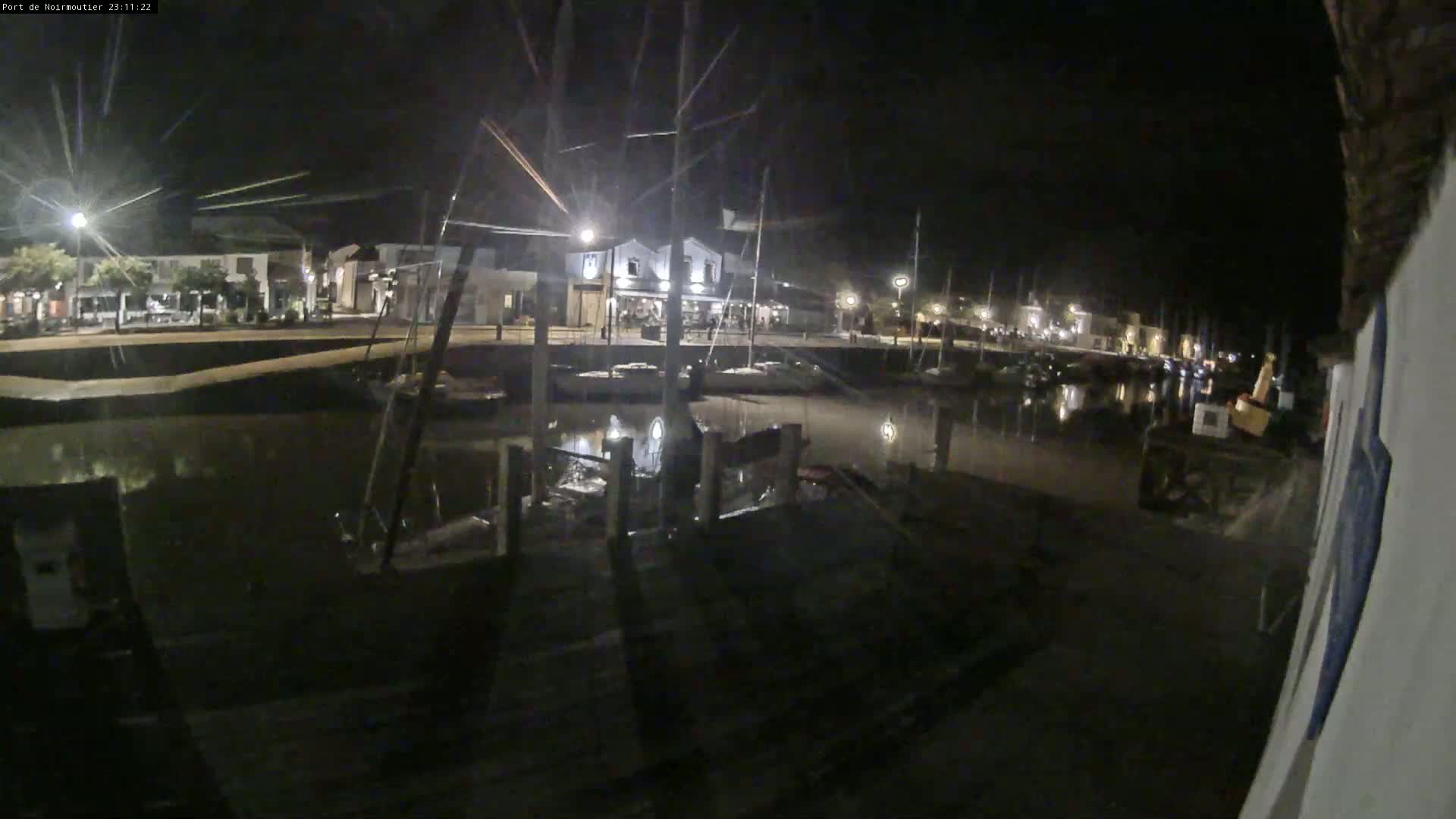 A nighttime view of a calm harbor with boats moored along a waterfront, buildings with lights visible across the water.