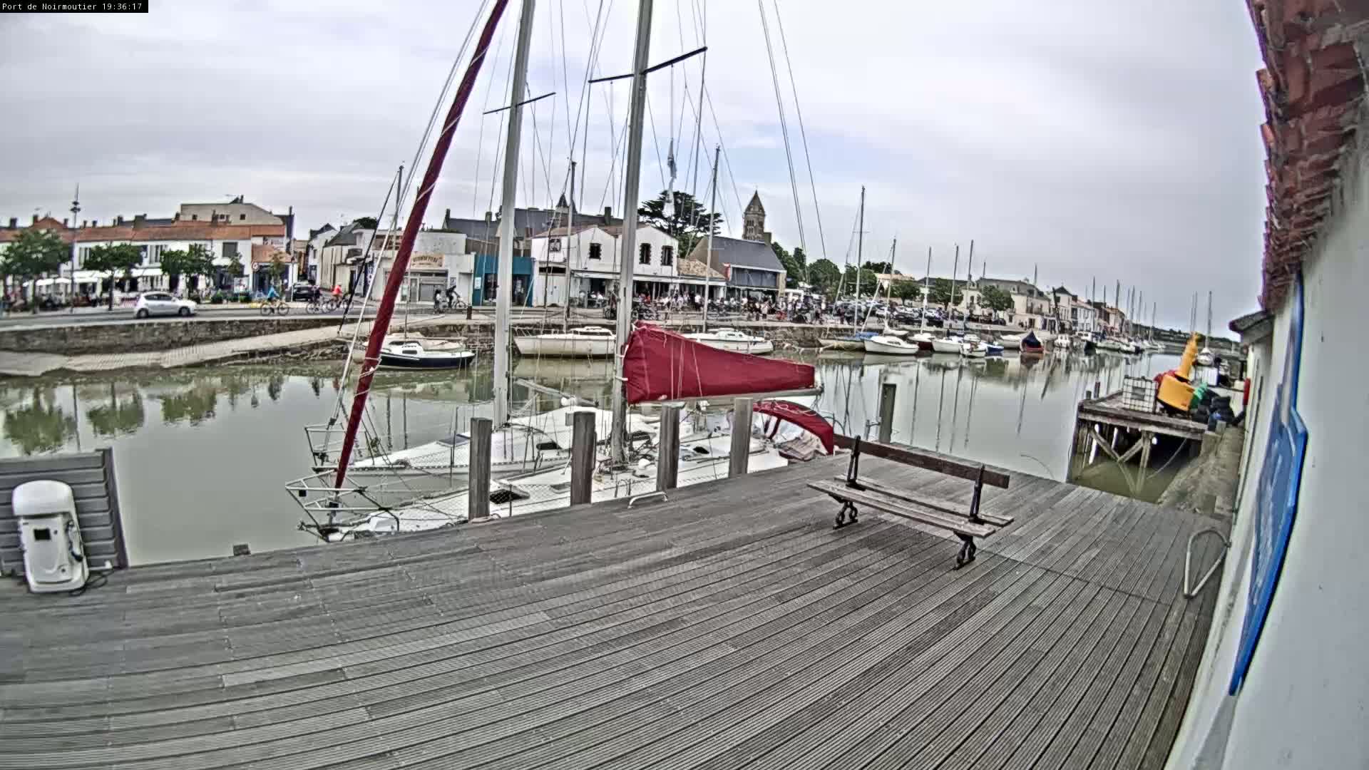 A wooden dock overlooking a calm harbor filled with sailboats, with a town visible in the background under an overcast sky.