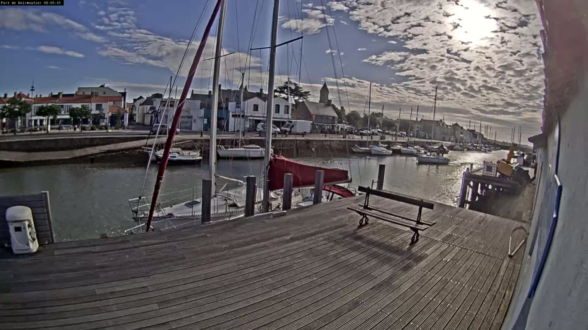 A partly sunny day at a harbor shows sailboats moored at a dock, with a wooden boardwalk and a bench in the foreground.