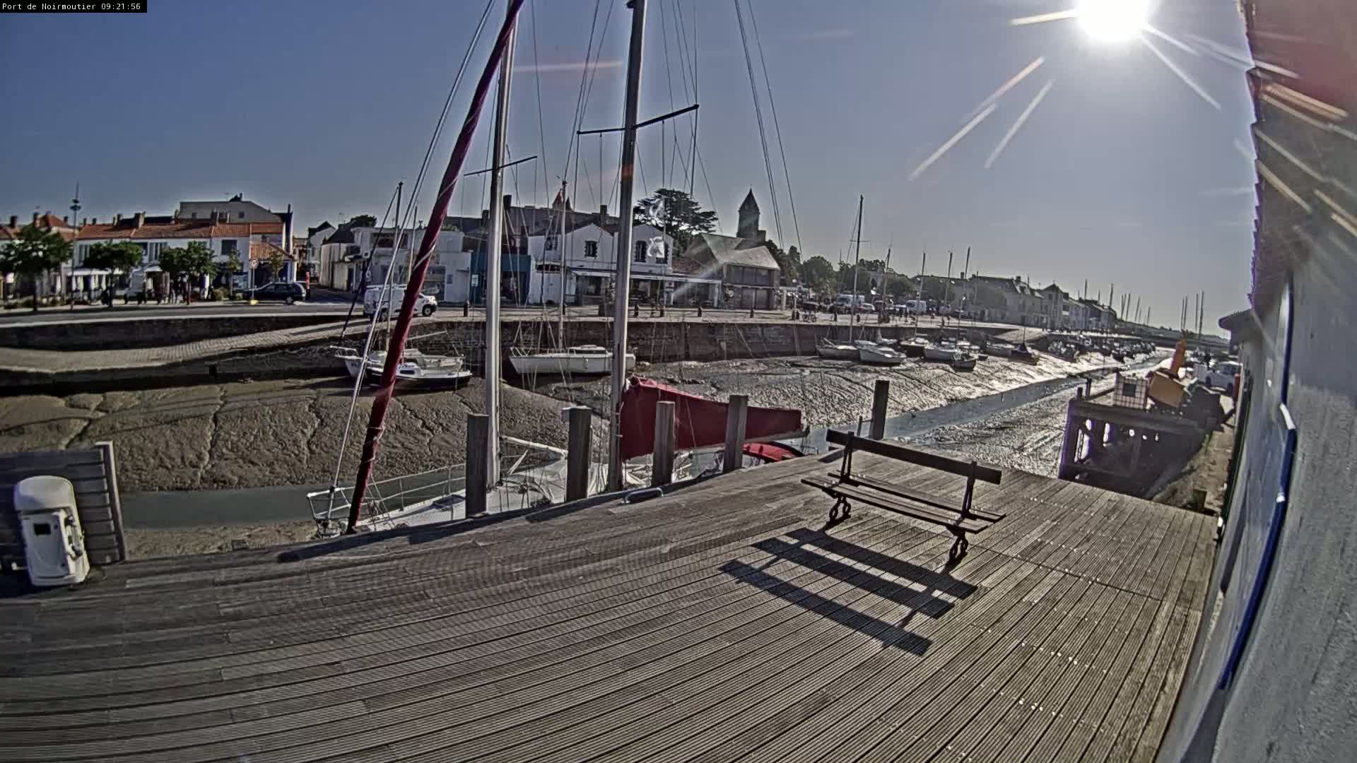 A sunny day at a harbor shows sailboats docked at low tide, with a wooden dock and bench in the foreground.
