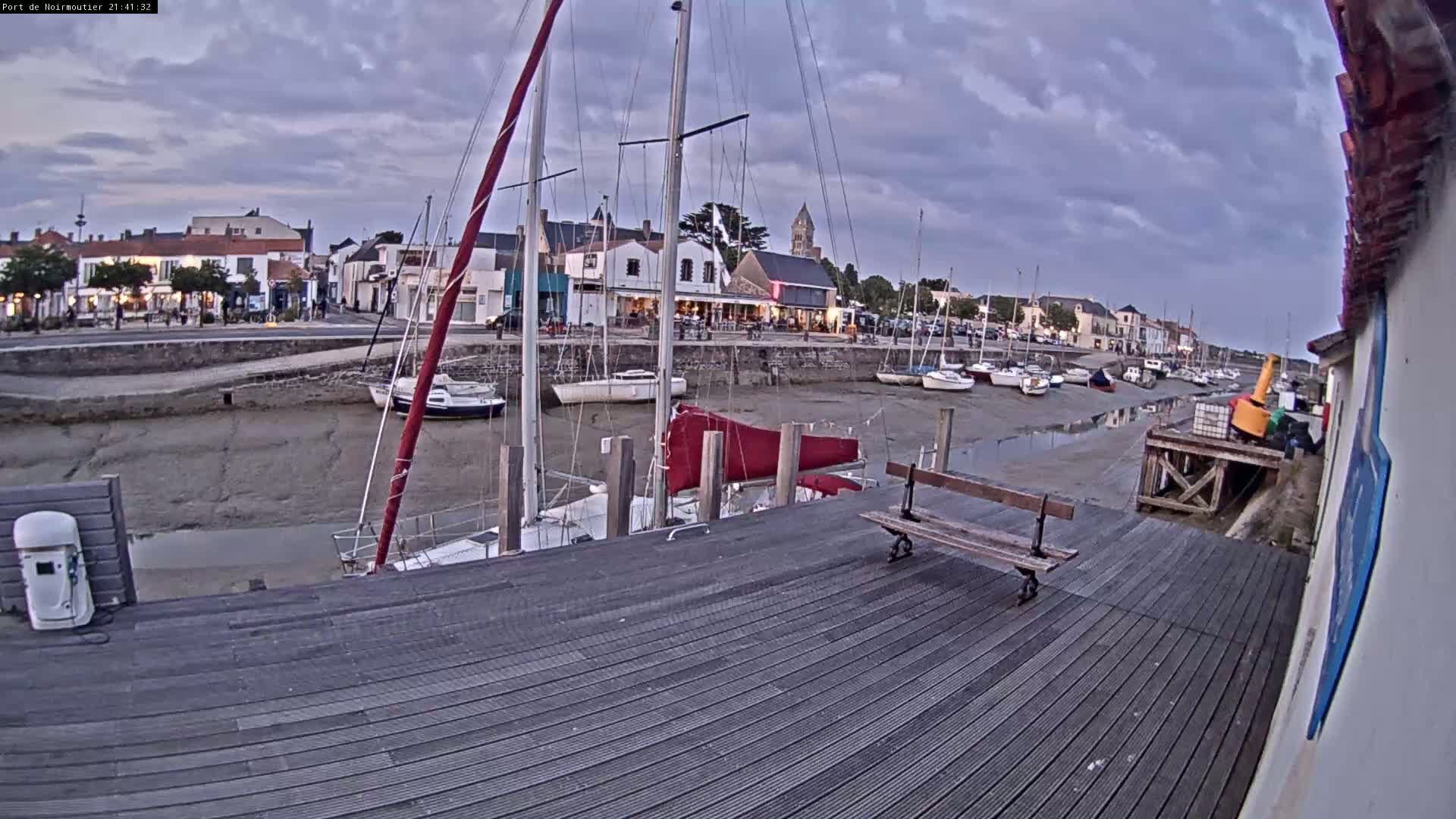 A wooden dock extends into a harbor at low tide, where sailboats are moored near a small town under a cloudy evening sky.