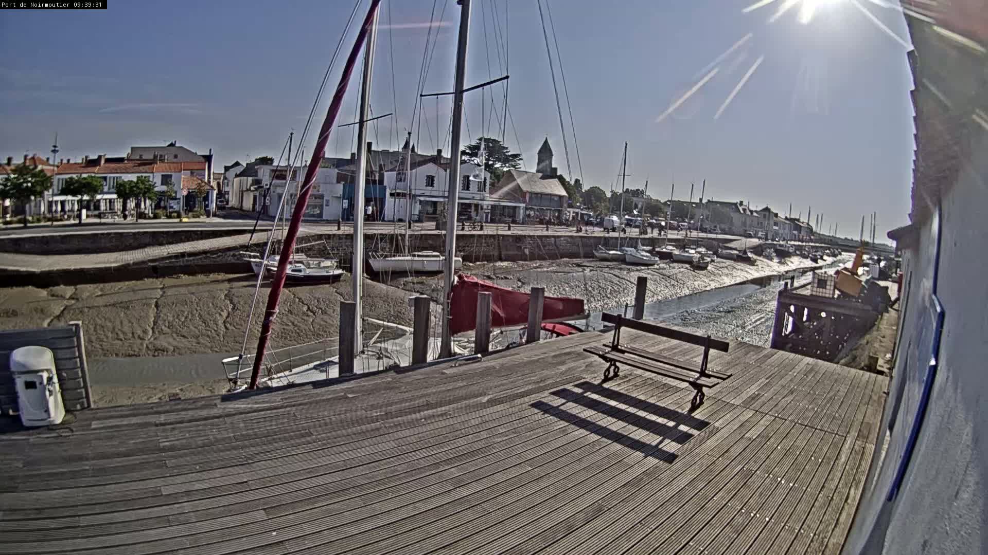 A sunny day at a harbor shows sailboats docked at low tide, with a wooden dock and bench in the foreground.