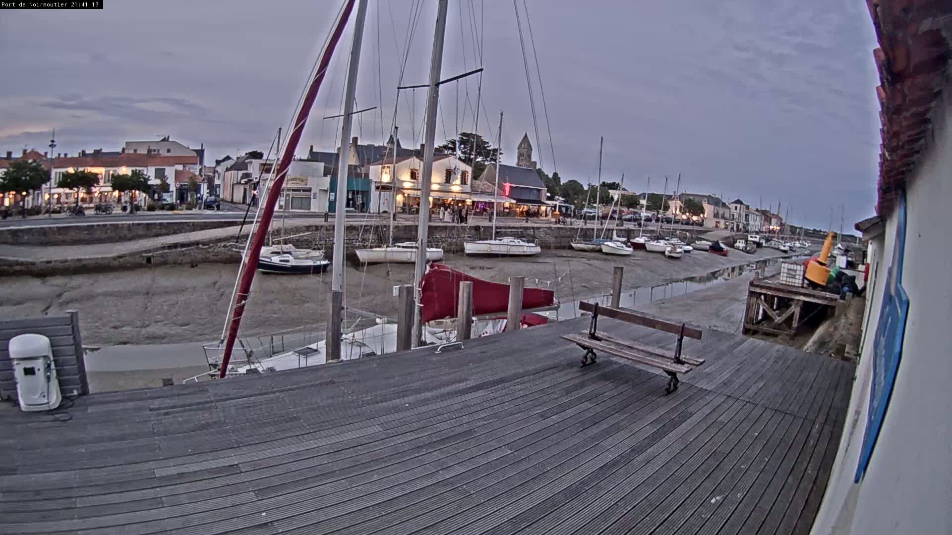 A harbor with numerous sailboats at low tide under an overcast sky, viewed from a wooden dock with a bench.