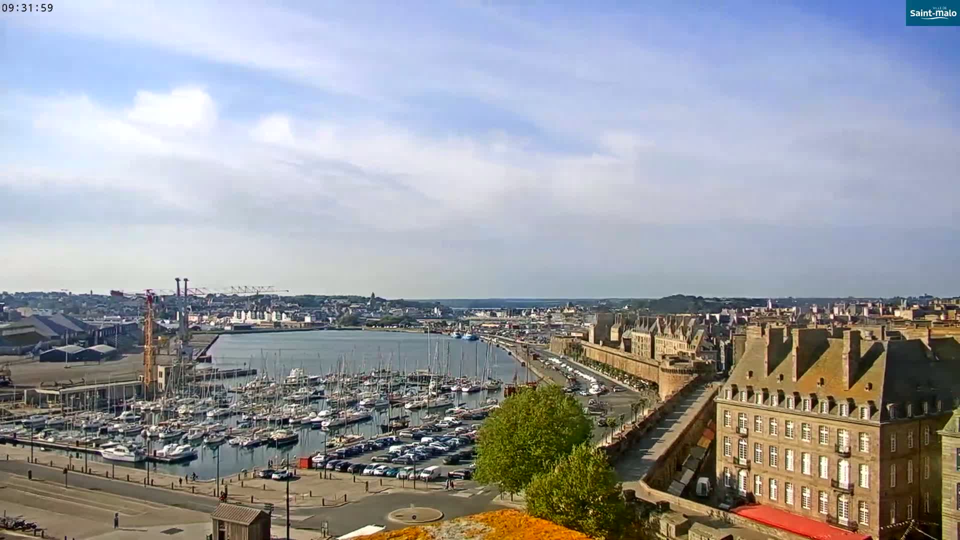 A sunny day reveals a harbor filled with numerous sailboats and yachts, adjacent to a city with historic stone buildings under a partly cloudy sky.