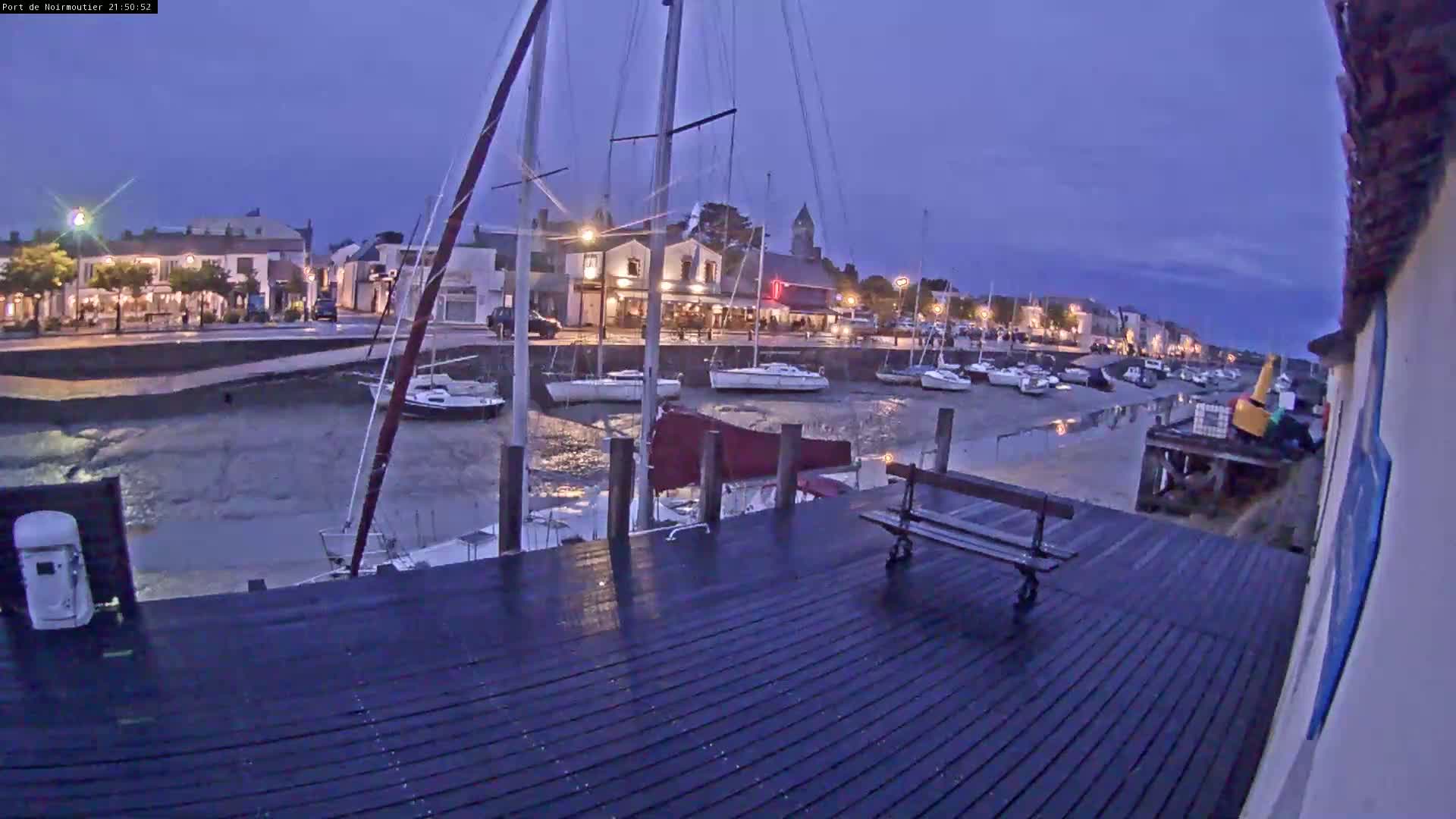 A dark, wet wooden dock at twilight overlooks a calm harbor filled with sailboats and a town lit by streetlights.