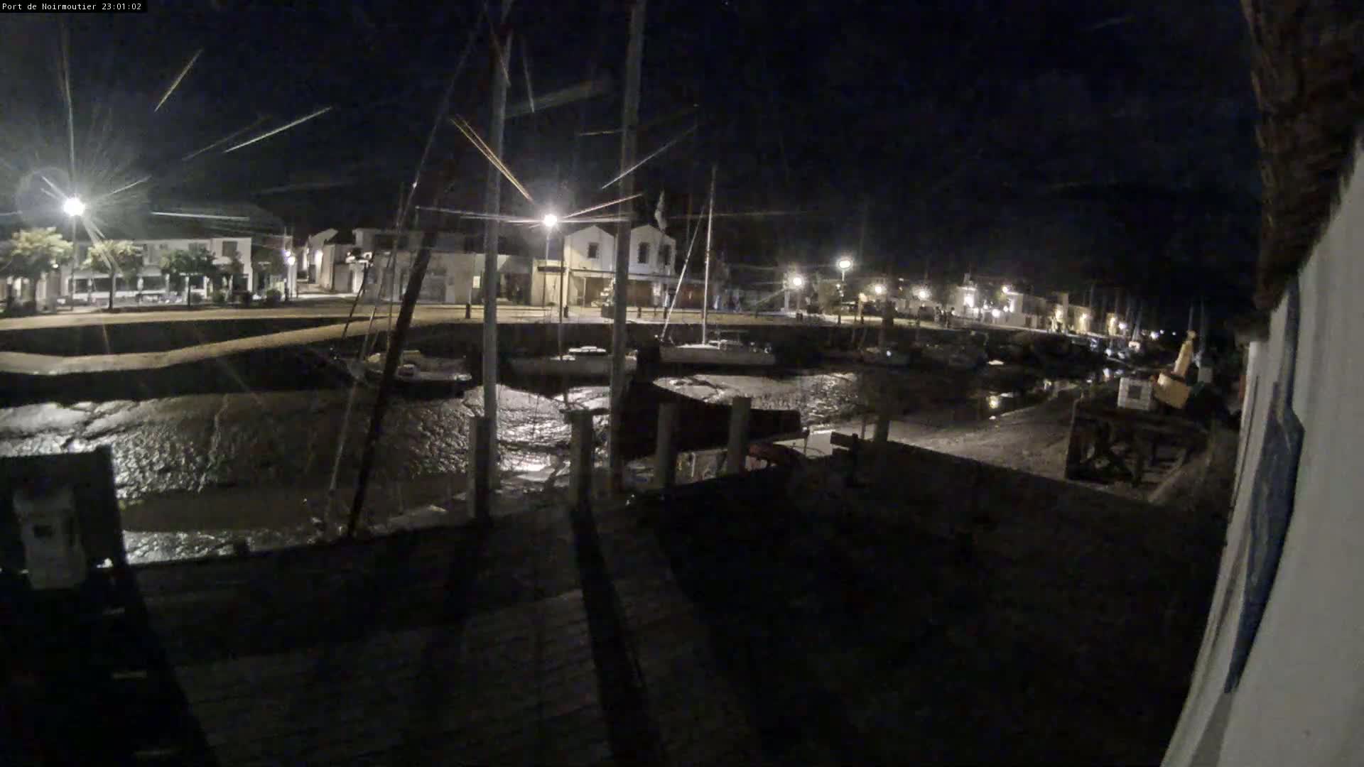 A nighttime view of a harbor with boats docked, low tide exposing a muddy seabed, and illuminated buildings lining the waterfront under a dark sky.