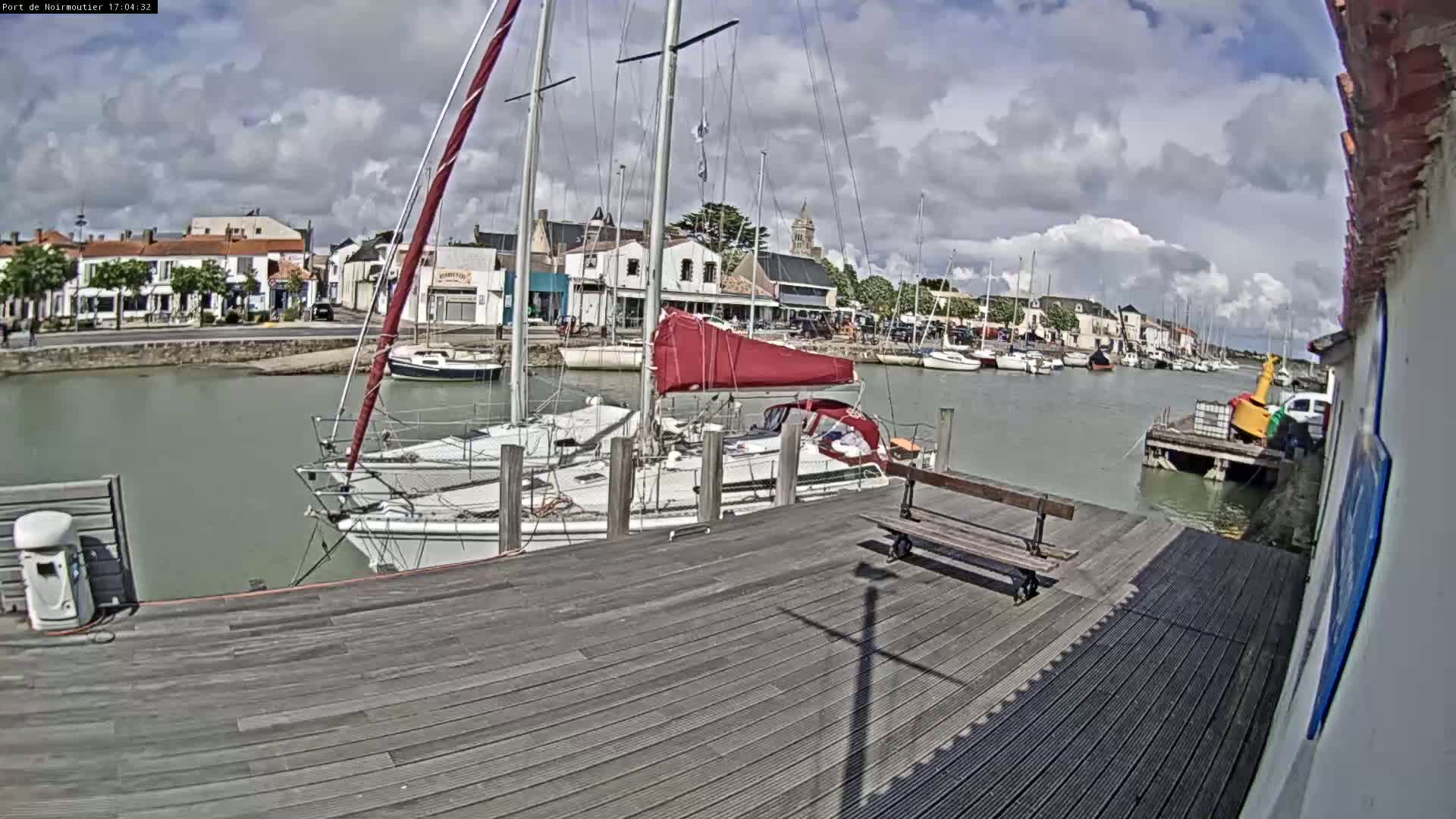 A wooden dock with several sailboats moored alongside it, overlooking a calm harbor with a village and partly cloudy sky in the background.