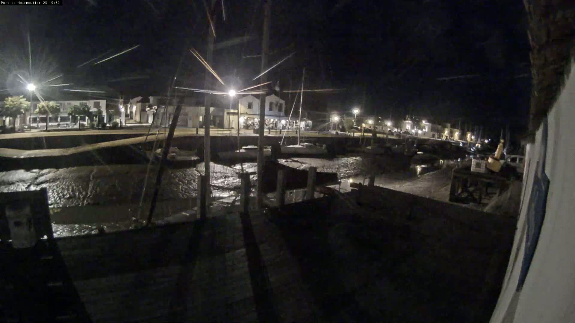 A nighttime view of a harbor with boats docked, low tide exposing muddy areas, and buildings with lights illuminated along the waterfront.