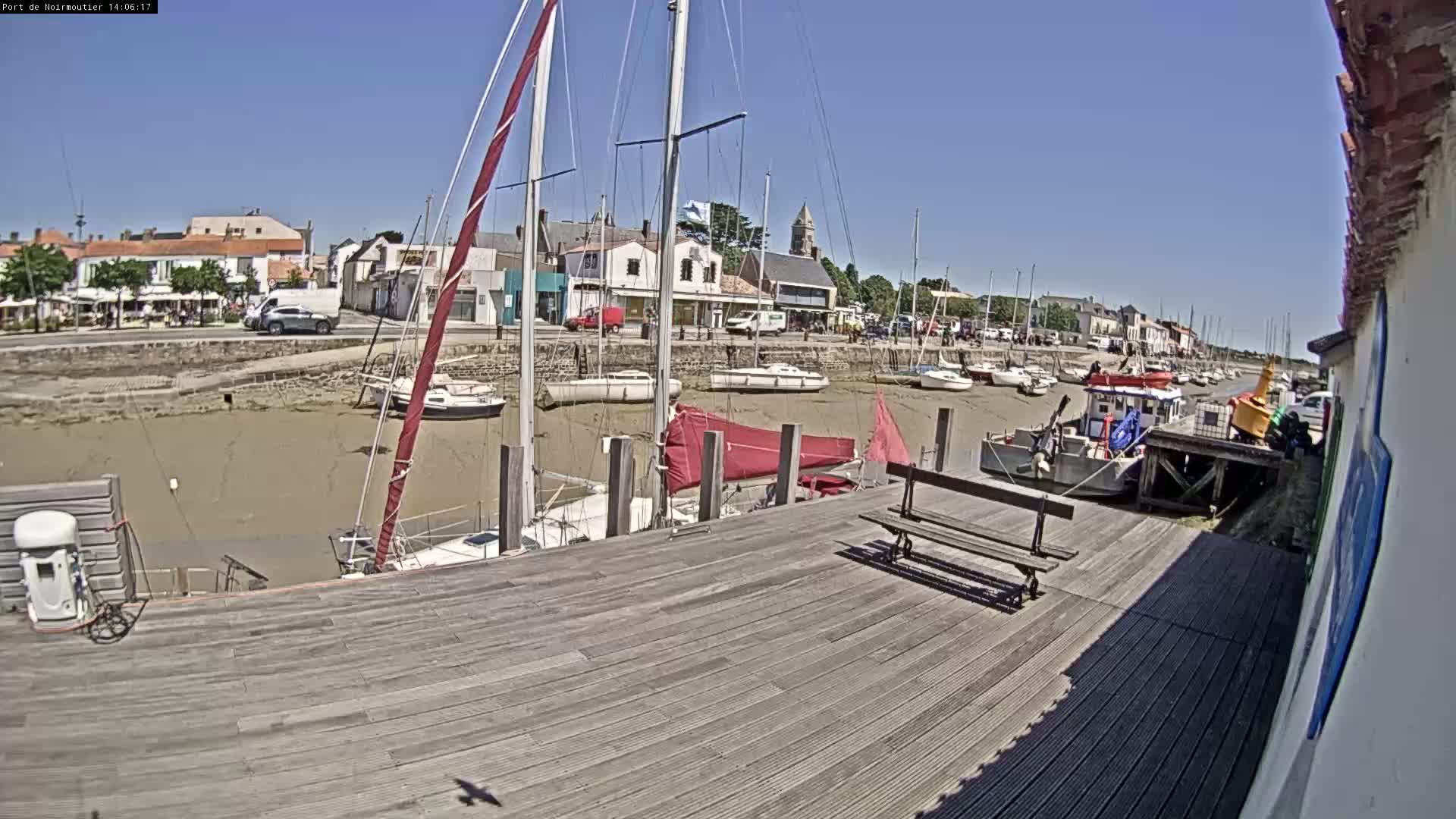 A sunny day at a harbor shows sailboats docked at low tide, with a wooden dock and a small town in the background.