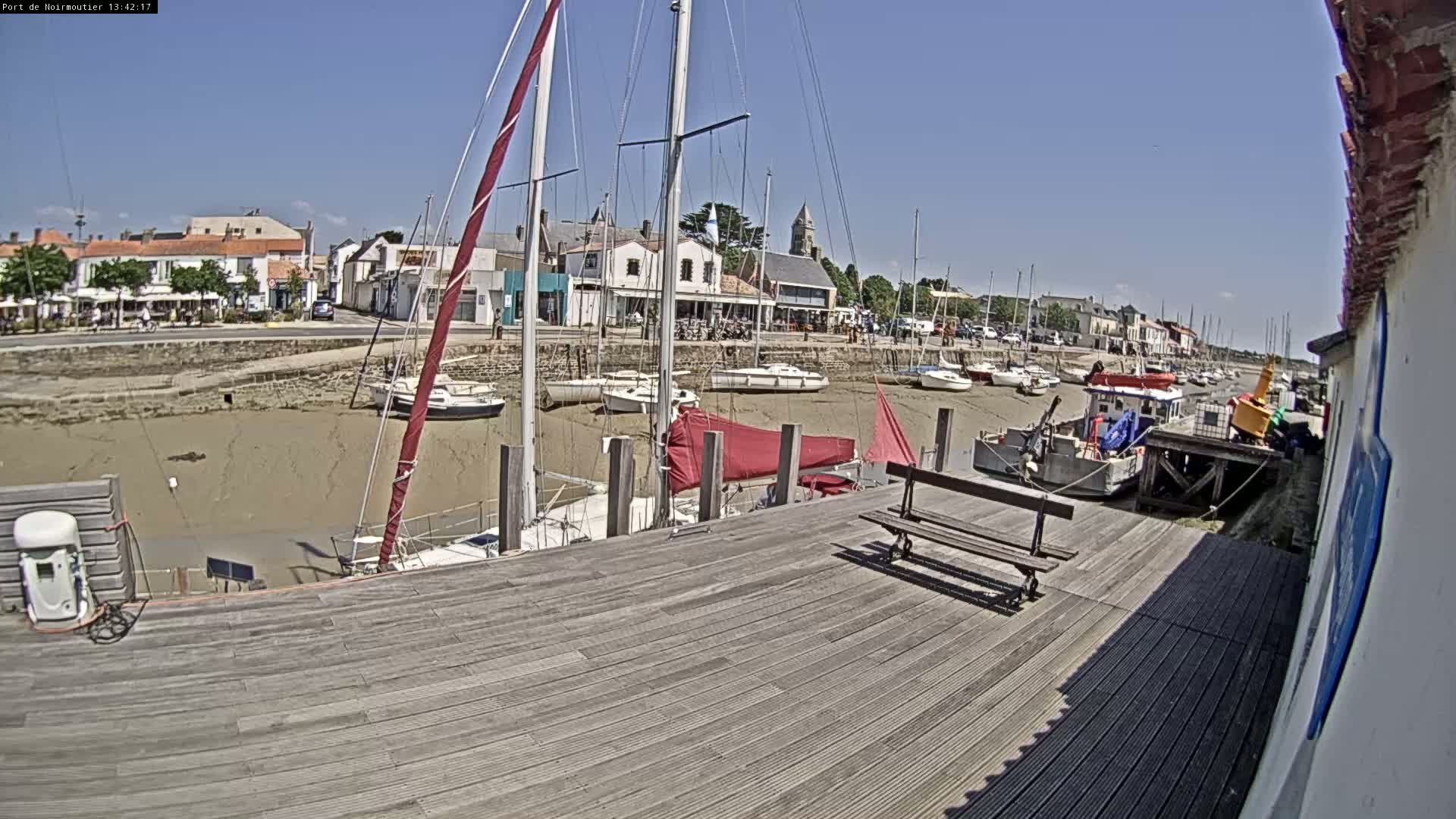 A sunny day at a harbor shows sailboats docked at low tide, with a wooden dock and a small town in the background.