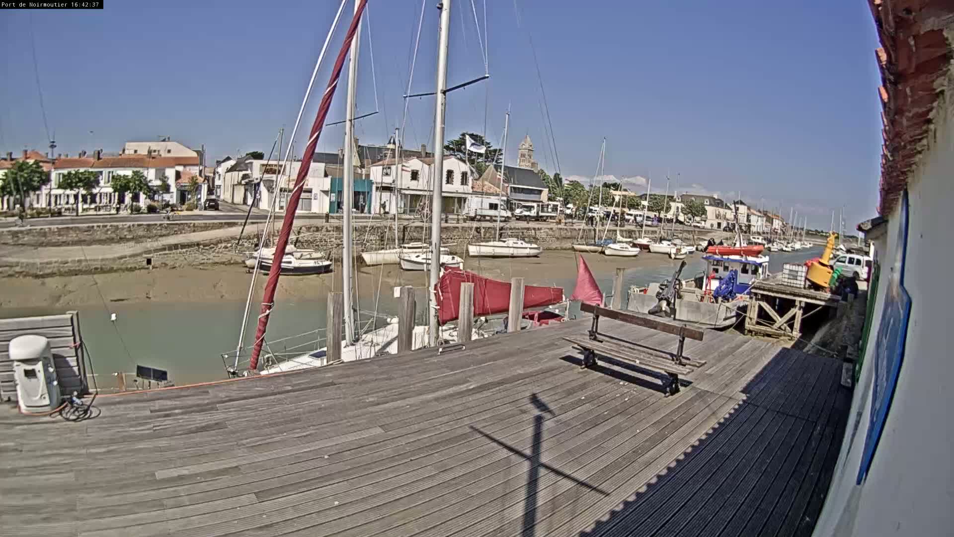 A sunny day at a harbor shows several sailboats docked along a wooden pier with a town visible in the background.