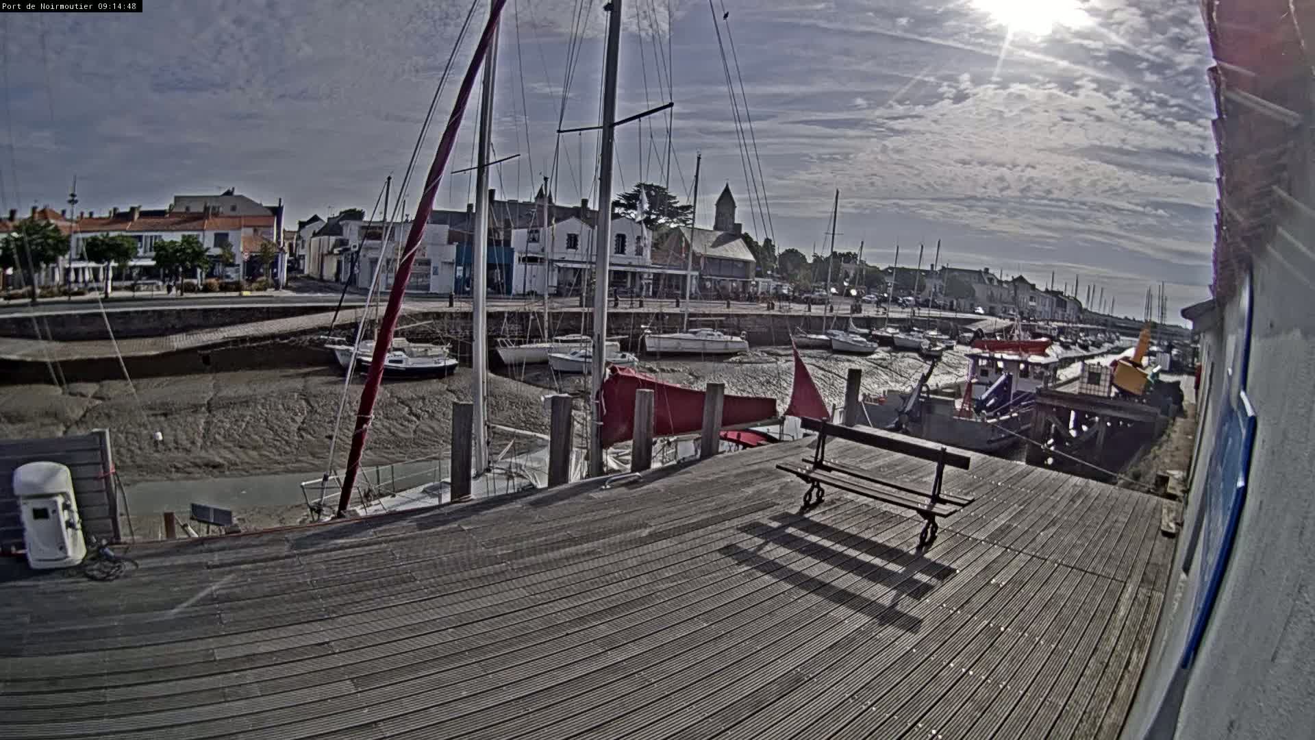 A mostly sunny day shows a harbor with sailboats docked, low tide exposing mudflats, and a wooden dock with a bench.