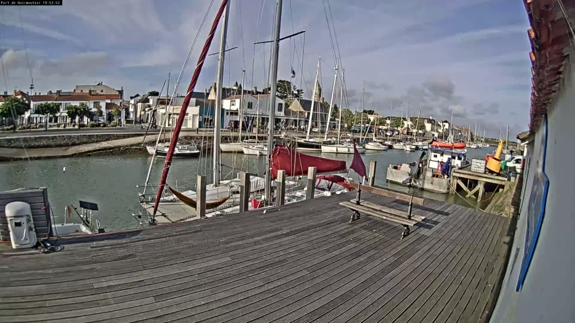 A wooden dock overlooks a calm harbor filled with sailboats and small motorboats under a partly cloudy sky.
