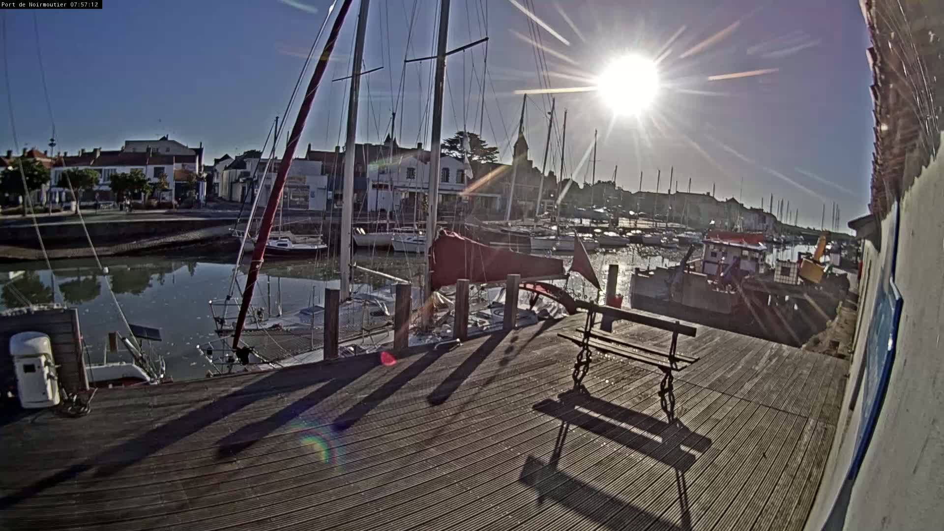 A sunny day at a harbor shows numerous sailboats docked along a wooden boardwalk with a bench and the shadows of posts cast on the planks.