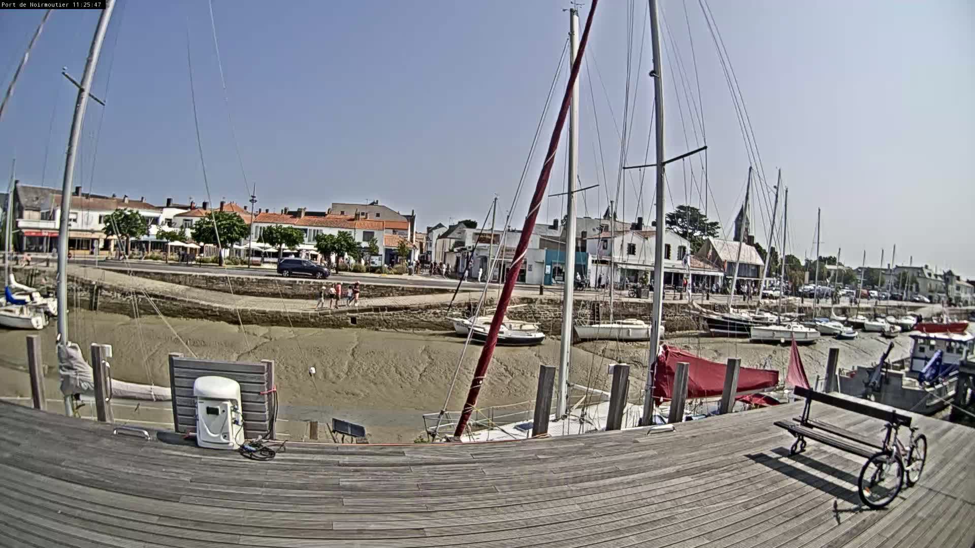 A sunny day at a harbor shows sailboats docked at low tide, with a wooden dock in the foreground and a village in the background.