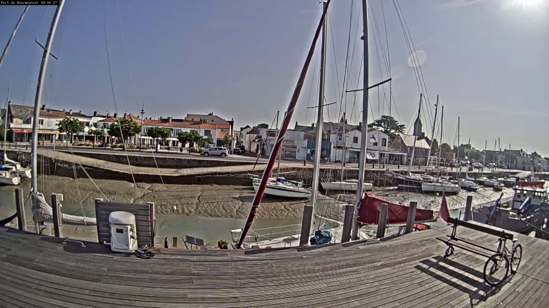 A sunny harbor scene shows sailboats docked at low tide, alongside a wooden boardwalk with a bench and bicycle.