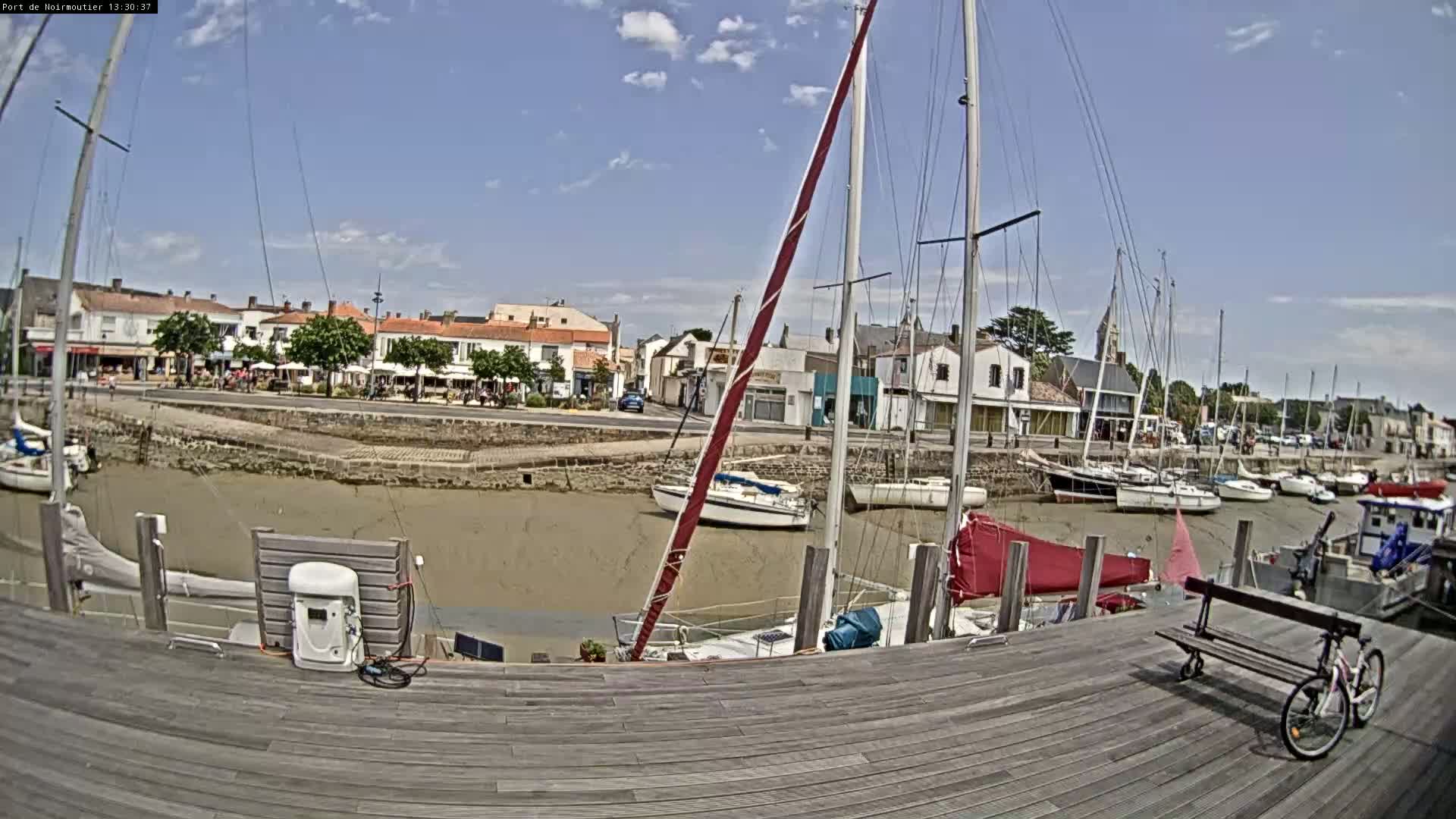 A harbor with sailboats docked, a low tide exposing muddy sand, and a town of buildings lining the waterfront under a mostly sunny sky.