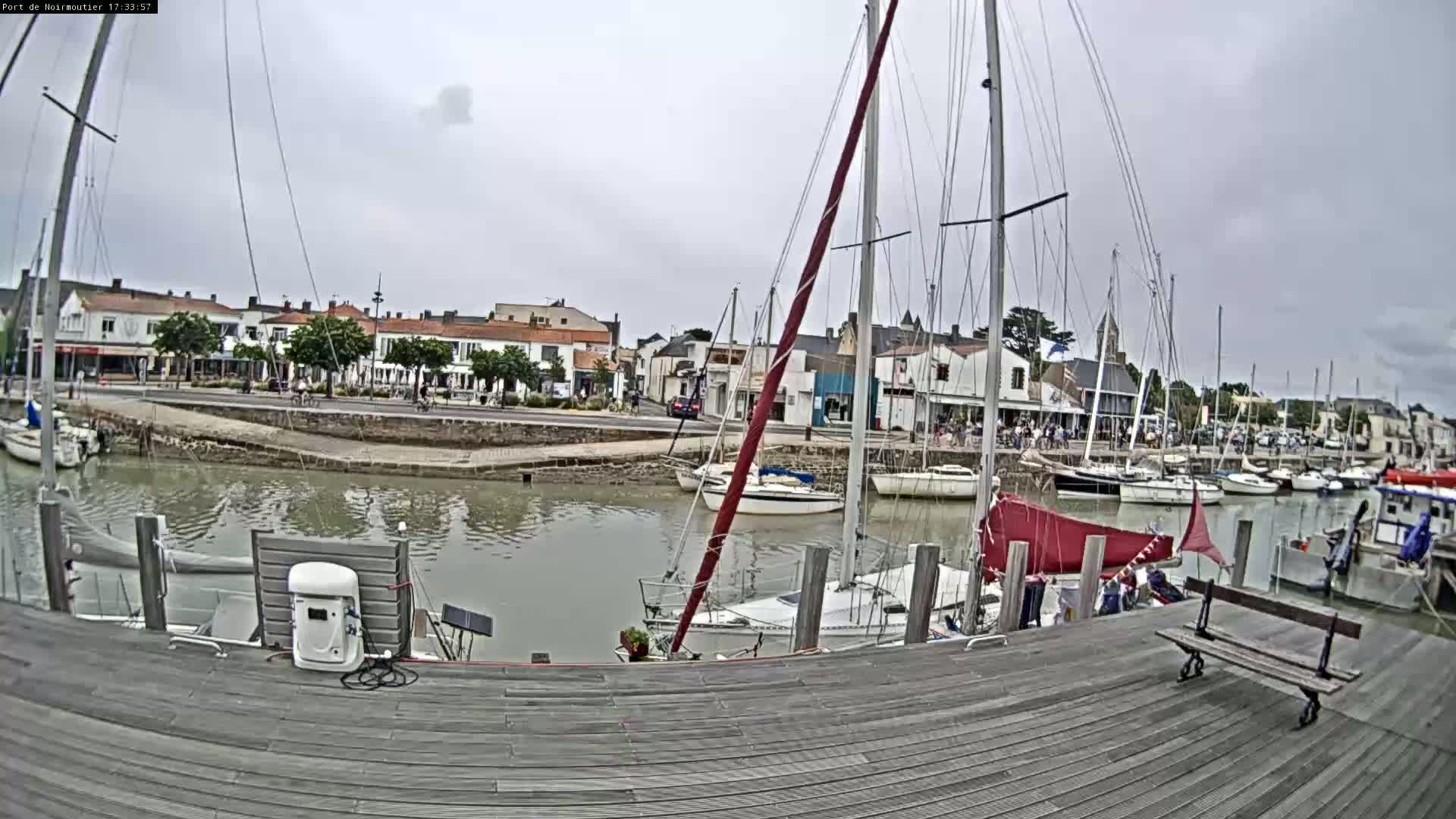 A harbor filled with sailboats under a cloudy sky, with a wooden dock in the foreground and buildings lining the waterfront.