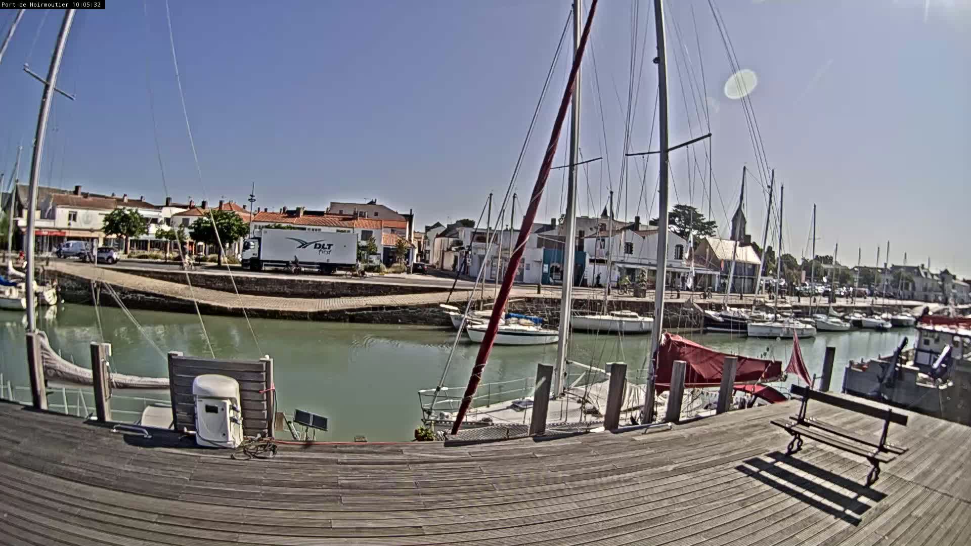 A sunny day at a harbor shows sailboats docked along a waterway next to a town with buildings and a paved area.
