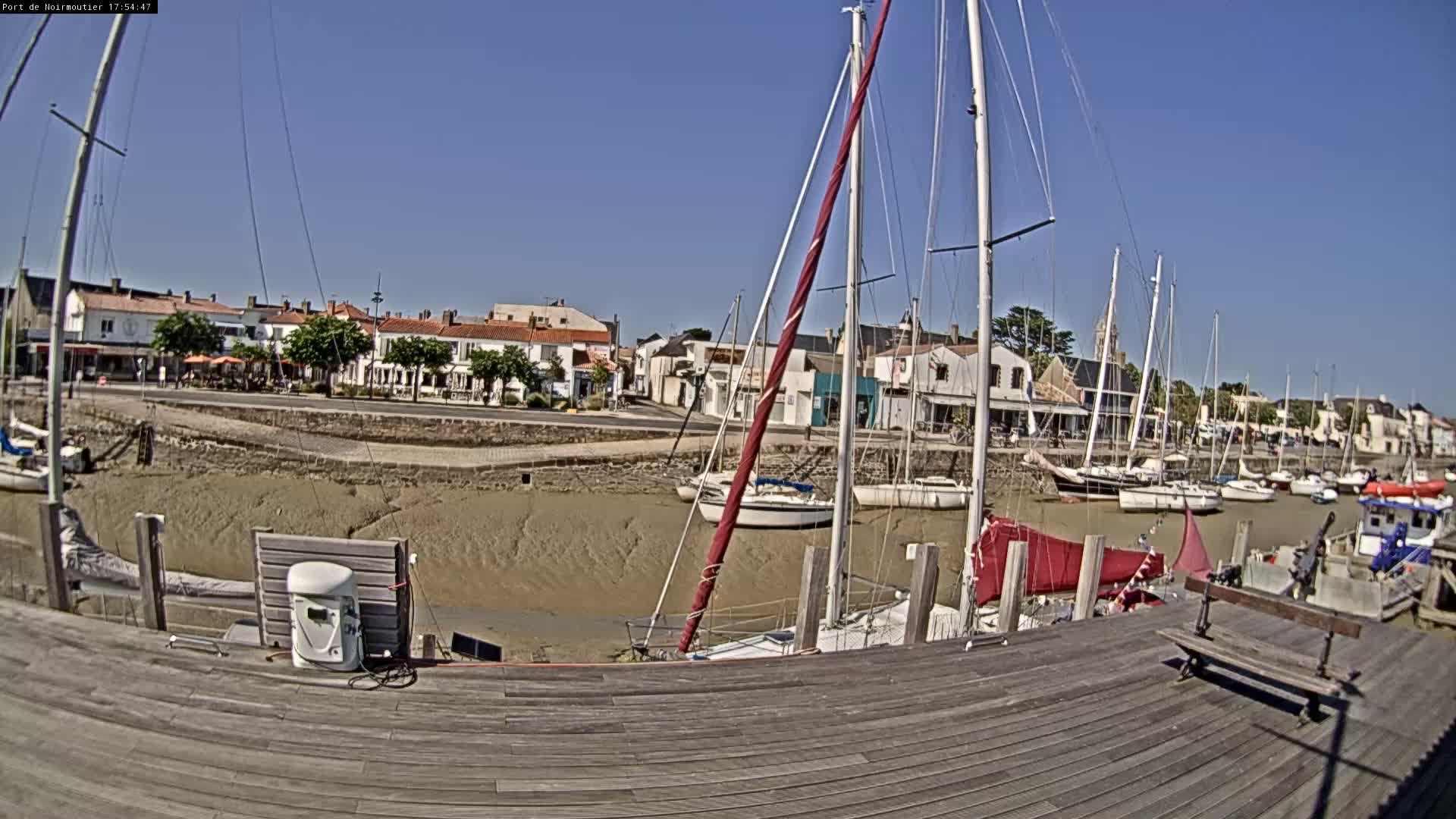 A sunny day at a harbor shows sailboats docked at low tide in front of a village with buildings lining the waterfront.