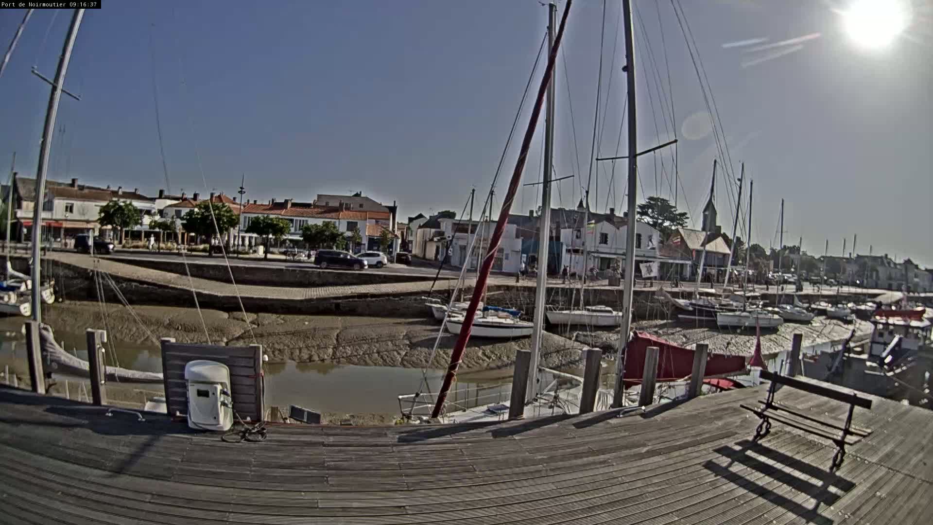 A sunny day shows a harbor with sailboats docked at low tide, a wooden dock in the foreground, and buildings lining the waterfront.