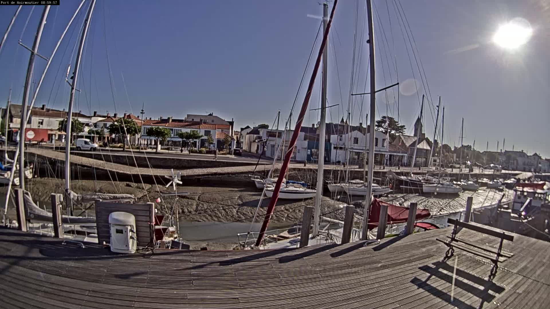 A sunny day at a harbor shows sailboats docked at low tide, with a village in the background.