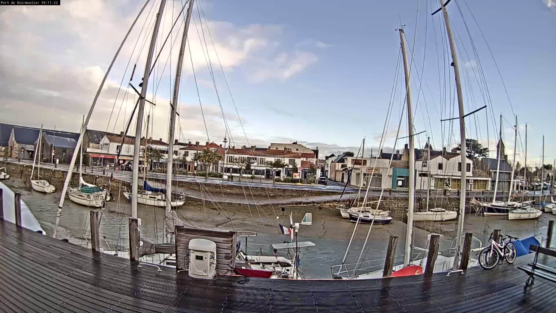Numerous sailboats rest on a muddy seabed in a partially drained harbor during low tide, overlooked by a coastal town under a partly cloudy sky.