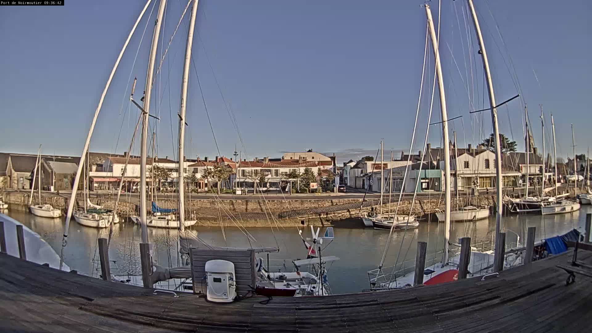 Numerous sailboats are docked in a calm, narrow waterway lined with waterfront buildings under a clear blue, sunny sky, viewed from a wooden pier in the foreground.