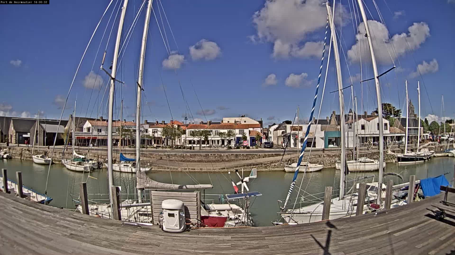 Numerous sailboats are docked in a calm, narrow waterway lined with waterfront buildings under a clear blue, sunny sky, viewed from a wooden pier in the foreground.