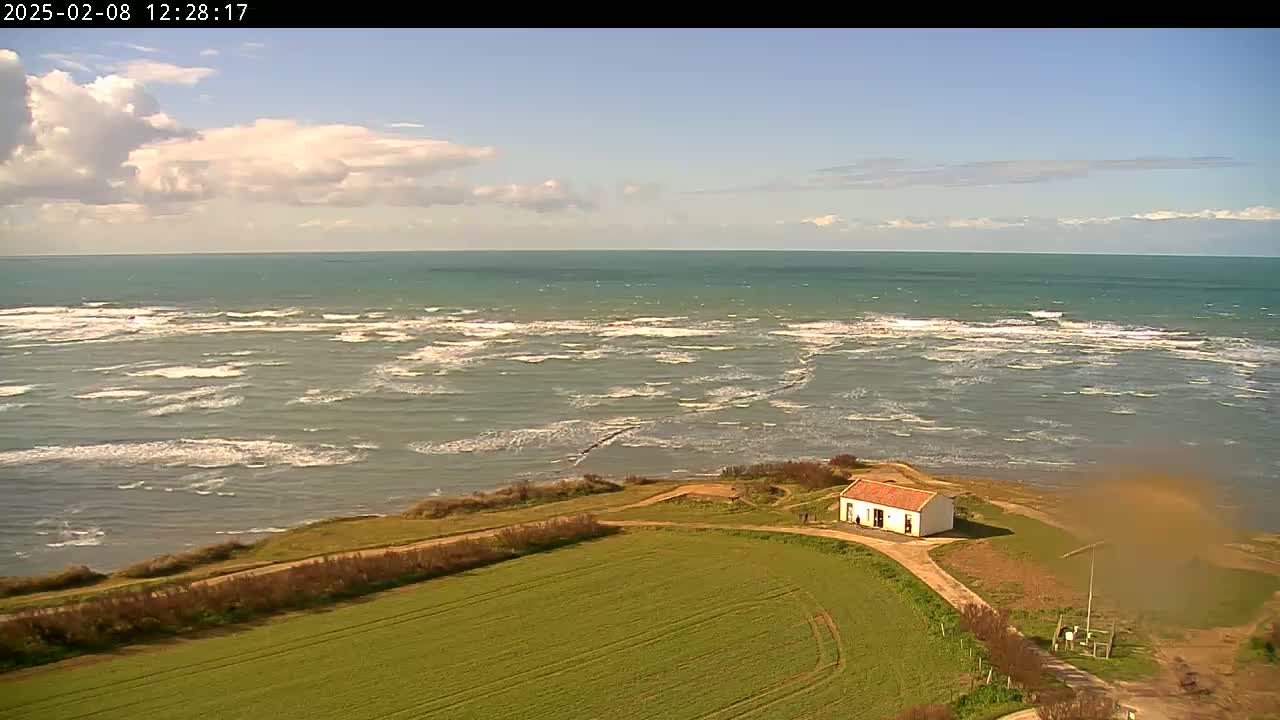 A small white building with a red roof sits on a grassy hill overlooking a choppy sea under a partly cloudy sky.