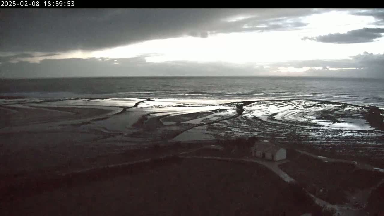 A dark, overcast sky hangs over a rocky, tide-pooled shoreline and a calm sea at dusk.