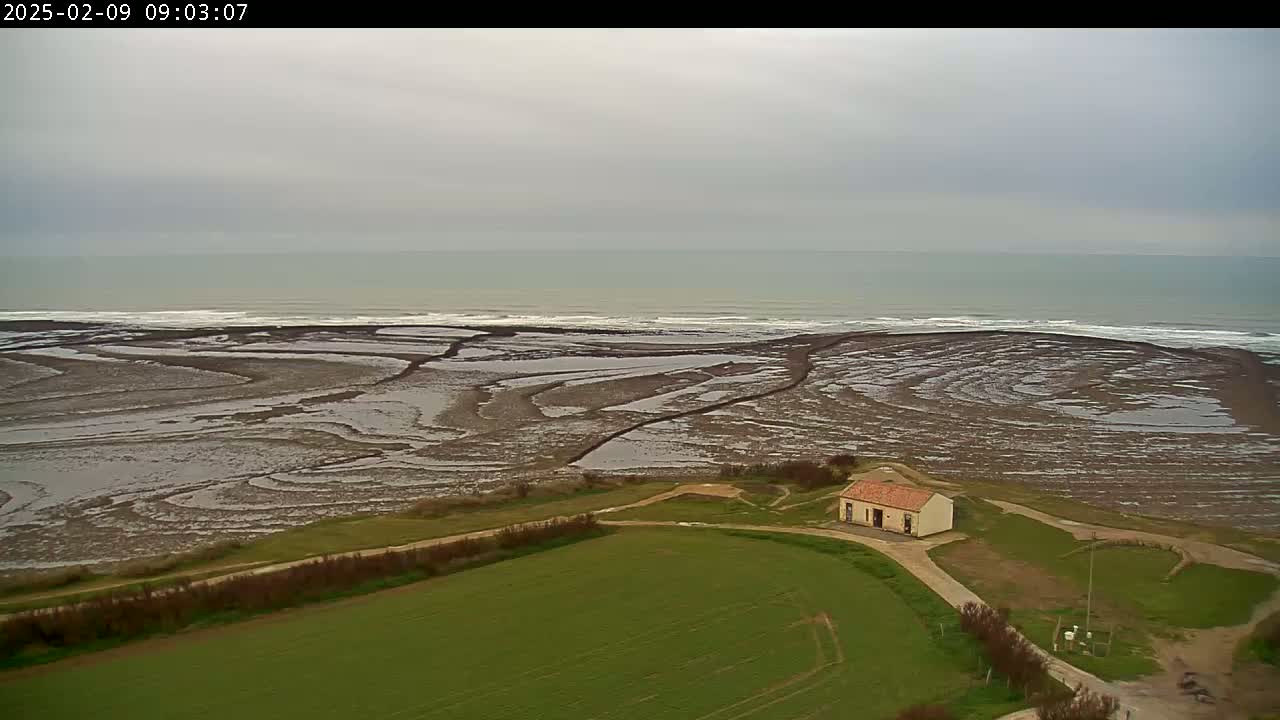 An overcast day reveals a small building situated on a grassy field overlooking a tidal flat that leads to a calm ocean.