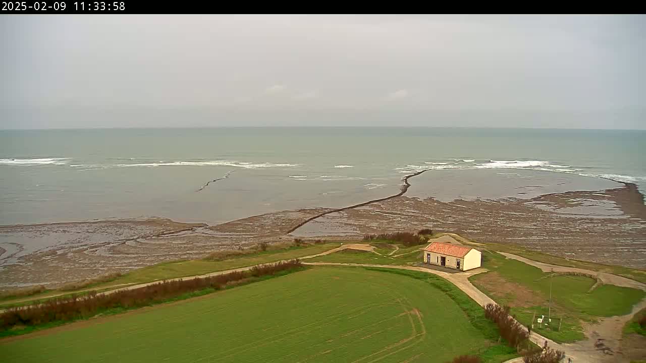 A small, light-colored building sits on a grassy hillside overlooking a calm, gray ocean with exposed, dark tidal flats and small waves under an overcast sky.