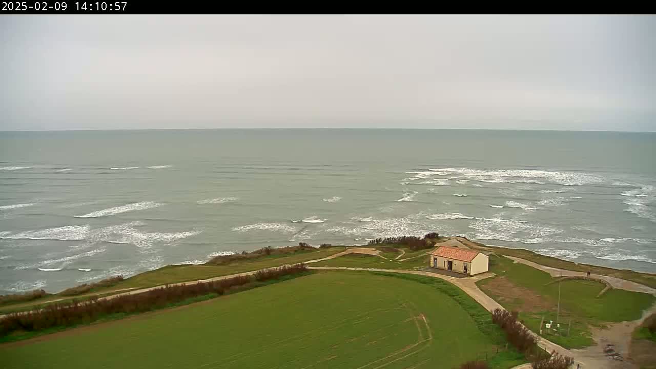 A small building sits on a grassy cliff overlooking a gray, wavy ocean under an overcast sky.