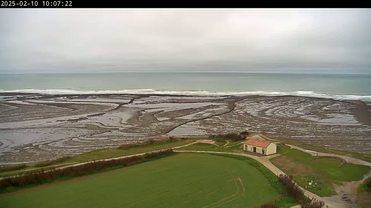An overcast day reveals a small building near a coast with a receding tide exposing intricate patterns in the mudflats, and a calm sea in the background.