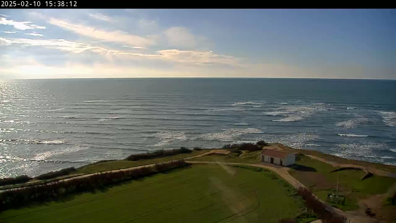 A small, white building sits on a grassy coastal area overlooking a wavy ocean under a partly cloudy sky.
