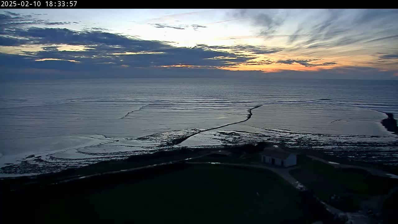 A coastal scene at sunset shows an expanse of ocean with exposed tidal flats extending towards a small building on a grassy area, under a partly cloudy sky.