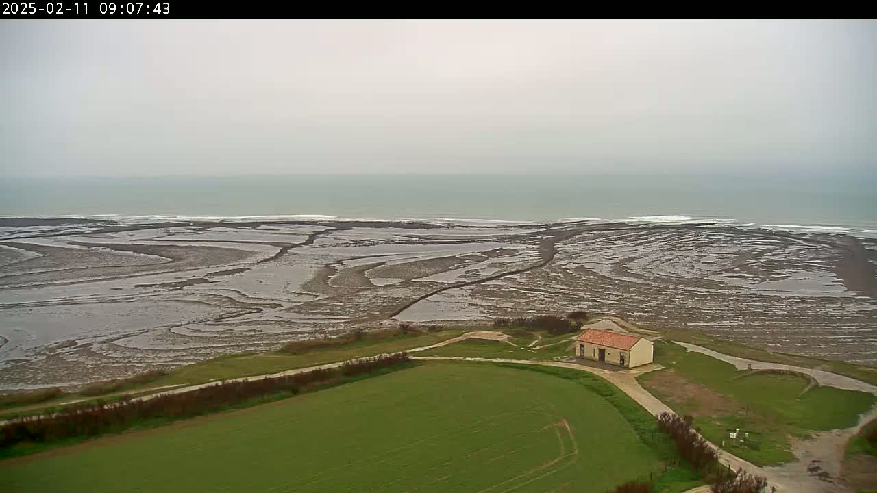 A small building sits on a grassy field overlooking a vast, mud-flat coastline and a grey, overcast ocean.