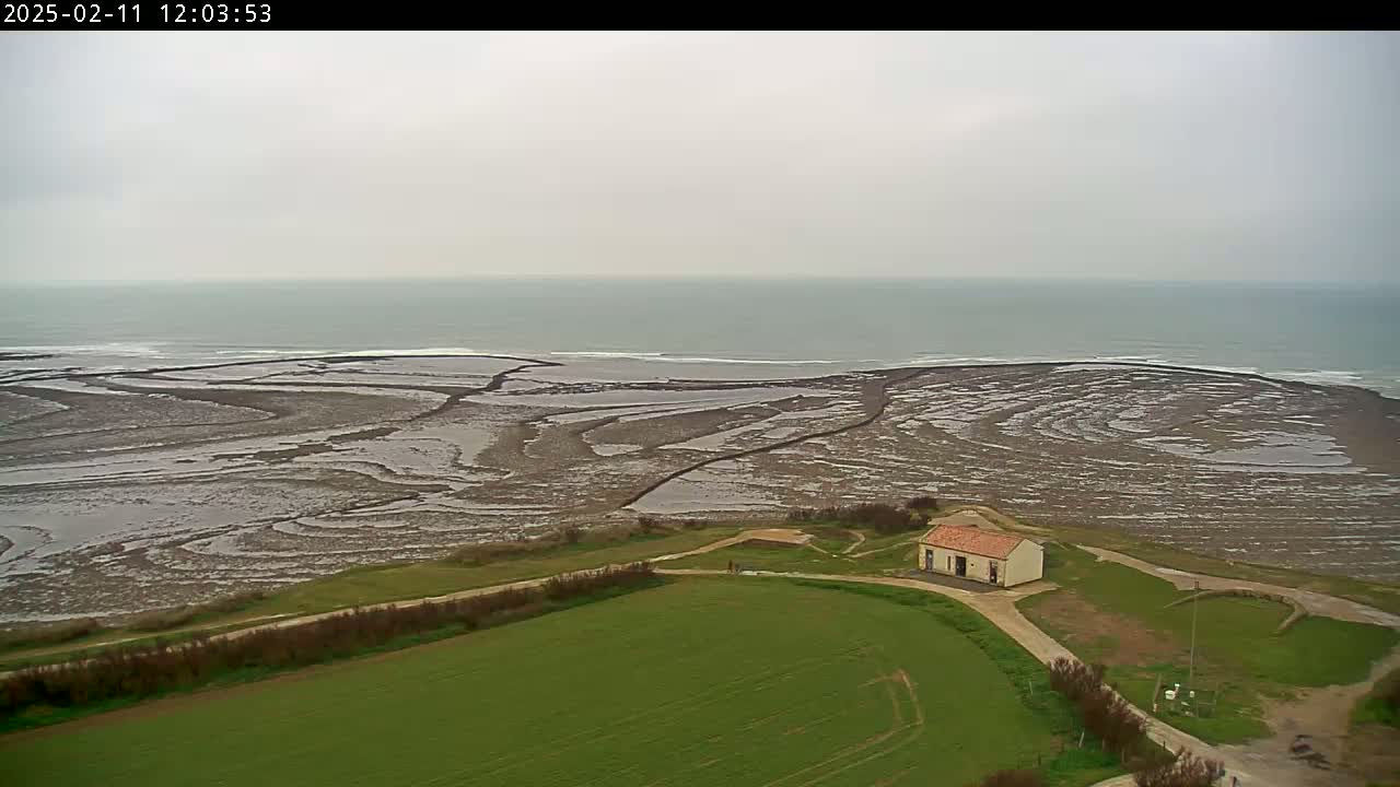 A small building sits on a grassy field overlooking a wide expanse of tidal flats leading to a calm, gray sea under an overcast sky.