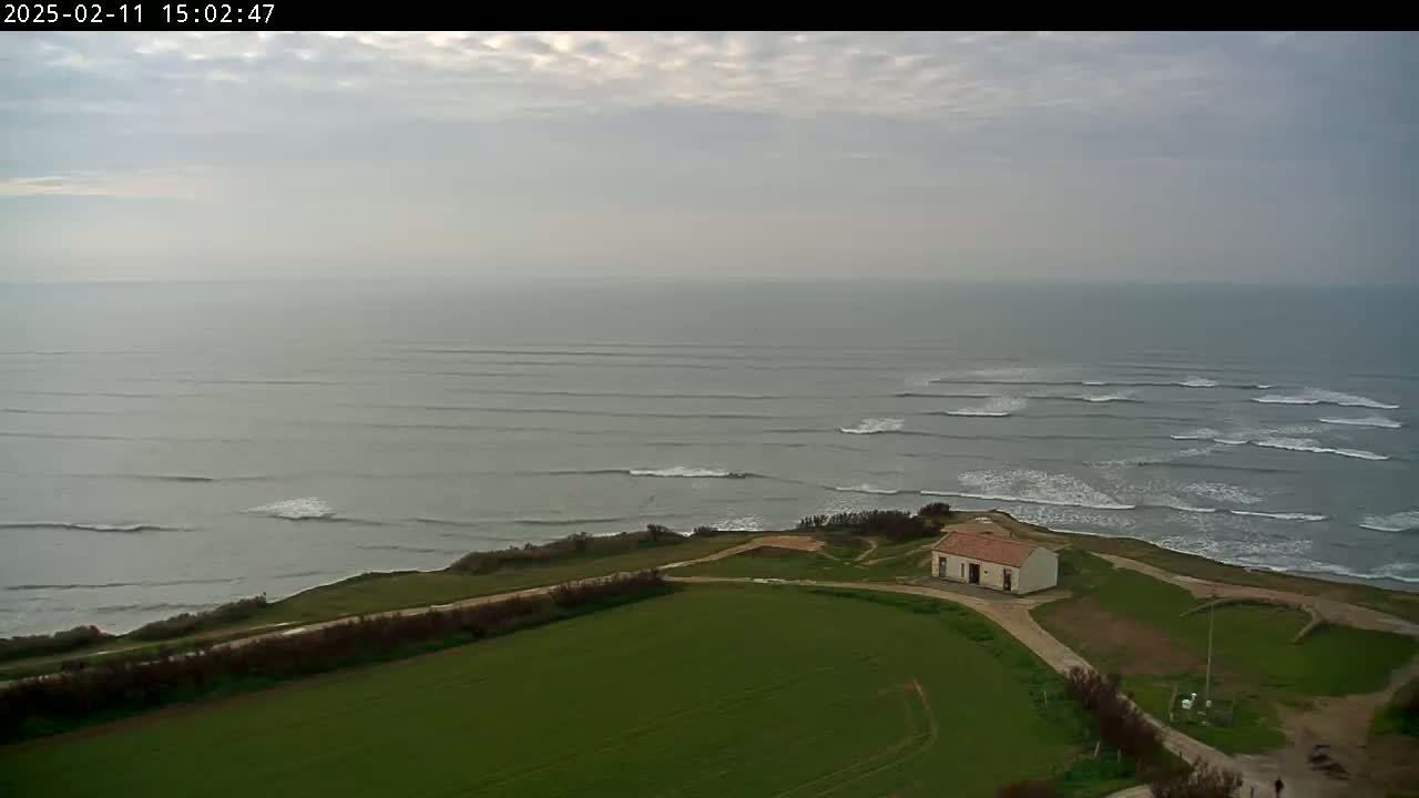 A small building sits on a grassy cliff overlooking a calm ocean with gently rolling waves under a cloudy sky.