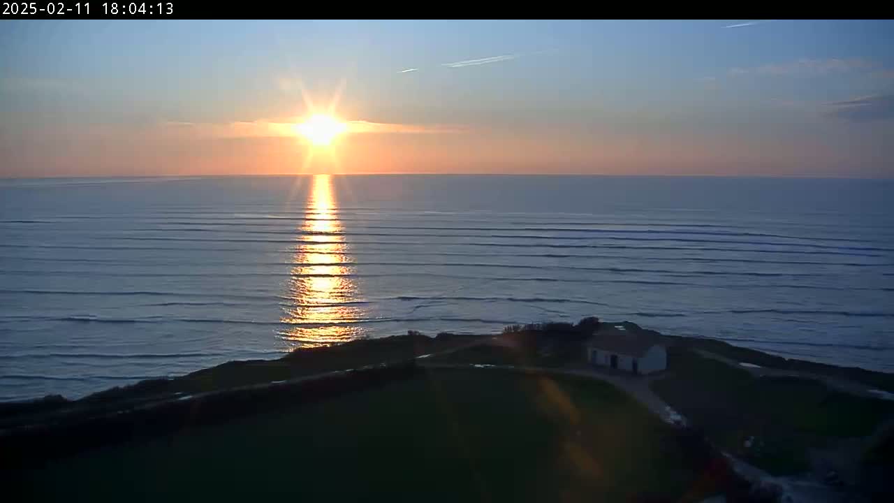 A sunset over a calm ocean with small waves is seen from an elevated vantage point, showing a small house on a grassy cliff overlooking the water.