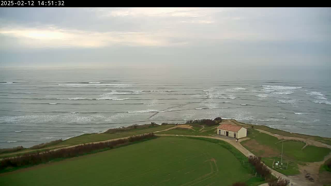 A small building sits on a grassy hillside overlooking a calm ocean with small, rolling waves under an overcast sky.