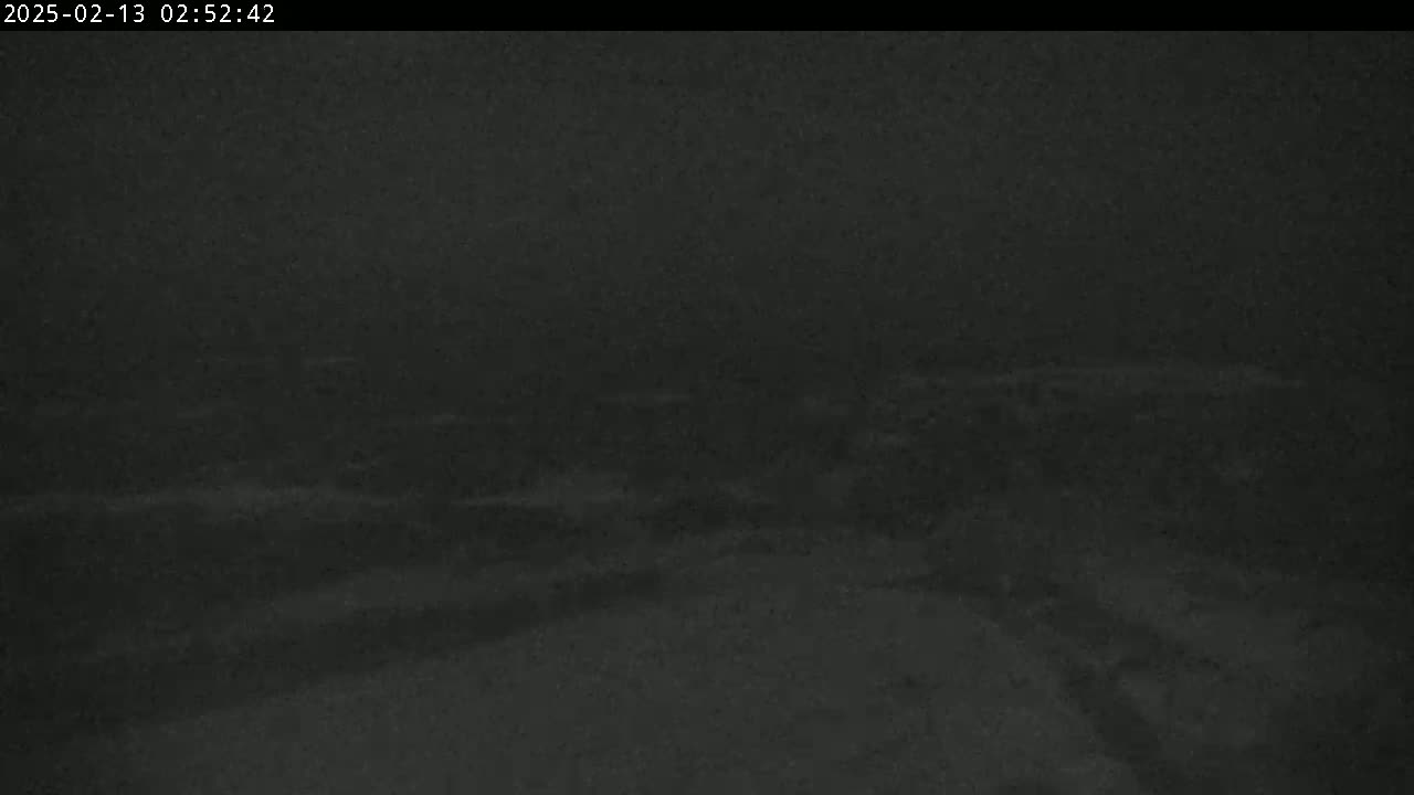 A low-light, nighttime outdoor scene shows a snow-covered ground with indistinct shapes and possibly roads in the distance under cloudy or snowy conditions.