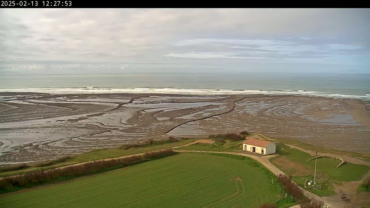 A small, light-colored building sits on a grassy field overlooking a wide expanse of exposed tidal flats and a gently waving ocean under a mostly cloudy sky.