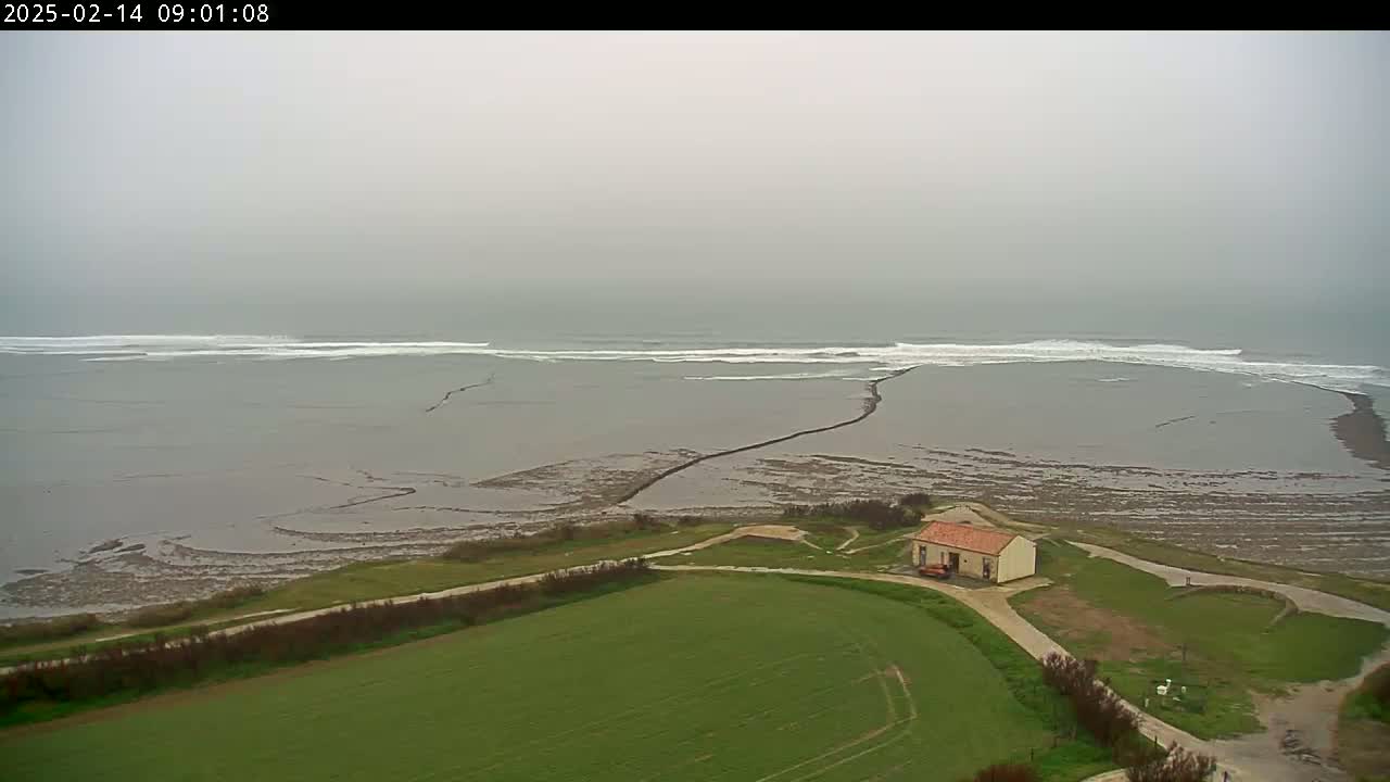 A small house sits on a grassy area overlooking a gray, overcast ocean with small waves breaking on a partially exposed, mud-flat shoreline.